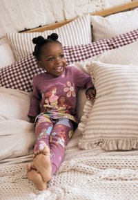 Lilla langærmede pyjamas med prinsegrafik og blomstermønster, kombineret med farverige leggings med mønster, sat på bløde puder.