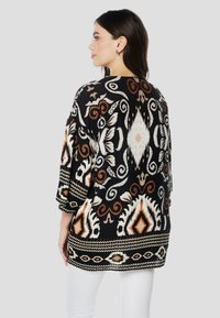 Cardigan negru cu modele albe, portocalii și bej. Prezintă mâneci largi și ornamente complexe de-a lungul tivului inferior.