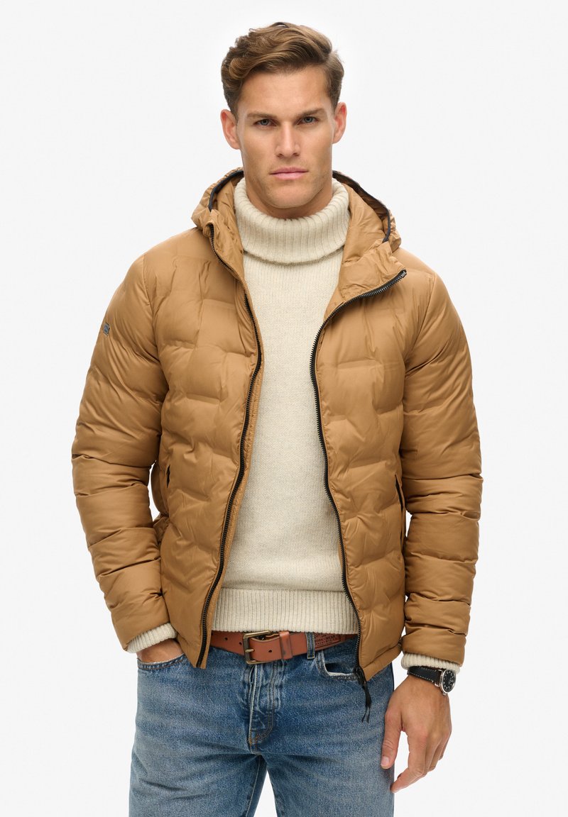 Bruine gewatteerde jas met capuchon, gestructureerde stof, met ritssluiting, gedragen over een crèmekleurige coltrui, gecombineerd met blauwe jeans en een bruine riem.