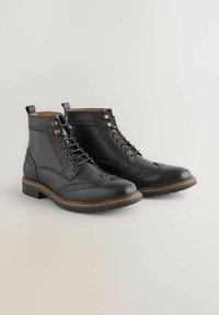 Čierne kožené brogove členkové topánky s wingtip detailom, šnurovacími prednými časťami a hnedo prešitou podrážkou na svetlom pozadí.