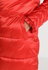 Rød polstret jakke lavet af shiny stof, med en sidelomme og quiltet tekstur med chevronmønstre på ærmerne.