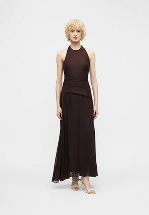 Ženska s kratkimi kodrastimi blond lasmi, oblečena v brezrokavno črno obleko z vrvjo okoli vratu in pliseiranim krilom ter bež sandalami z odprtimi prsti.