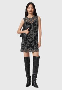 Zwart mouwloos jurkje met ingewikkelde zilveren bloemembroidery, met een geschulpte zoom, gecombineerd met zwarte hoge laarzen tot boven de knie.