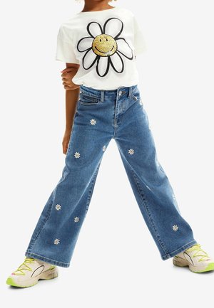 Camiseta blanca con una gran flor delineada en negro y lentejuelas doradas. Vaqueros de pierna ancha de denim azul adornados con bordado de margaritas blancas. Zapatillas con detalles verdes.