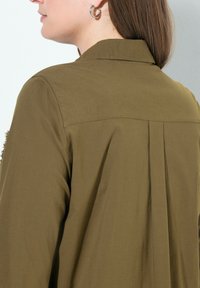 Camicia verde oliva con colletto, maniche lunghe e una piega centrale sul retro. Realizzata in tessuto morbido, presenta orecchini ad anello argentati.