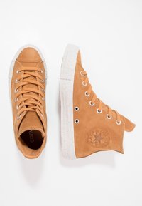 High-top sneakers in tan leather with a textured finish, white speckled rubber sole, and metal eyelets, featuring a logo patch on the side.