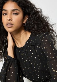 Schwarze Bluse mit goldenen Sternmustern, leichtem Stoff, gerafftem Ausschnitt, Schleifendetail und langen, transparenten Puffärmeln.