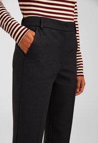 Pantaloni neri su misura con una finitura liscia e strutturata, dotati di tasche laterali e una piega frontale. Abbinati a una maglietta a righe con maniche lunghe.