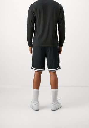 Personne debout de dos, portant un t-shirt noir à manches longues, un short noir avec des rayures blanches, des chaussettes blanches et des baskets blanches.