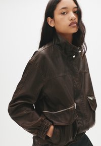 Giacca in finta pelle marrone con colletto alto, tasche con zip, polsini elasticizzati e una vestibilità rilassata, che presenta una texture liscia e cuciture minime.