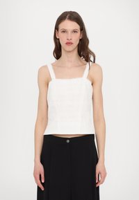 Top court blanc avec décolleté carré, fines bretelles réglables, texture brodée et accents à motifs horizontaux. Associé à un pantalon noir.