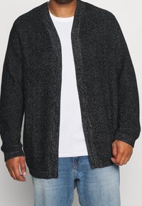 Svart stickad cardigan med en strukturerad yta, öppen framsida, långa ärmar och avslappnad passform, bärs över en vit t-shirt och jeans.