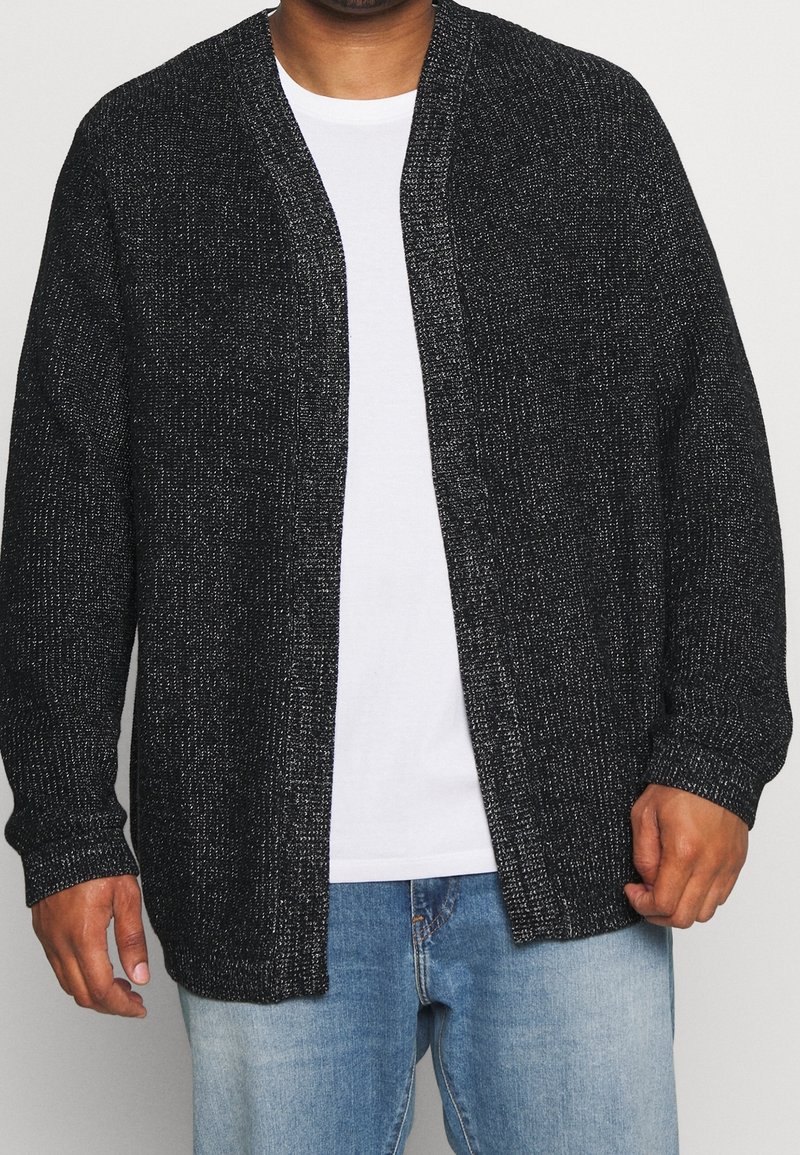 Svart stickad cardigan med en strukturerad yta, öppen framsida, långa ärmar och avslappnad passform, bärs över en vit t-shirt och jeans.