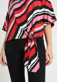 Blusa a righe rossa, rosa e nera con maniche corte, caratterizzata da un dettaglio a nodo in vita. Il materiale sembra morbido con una texture liscia.