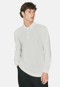 Ilgai rankovėmis polo marškiniai su tekstūruotu šviesiai balto audinio paviršiumi, turintys smailų apykaklę ir tris mygtukus, derinami su tamsiomis kelnėmis.