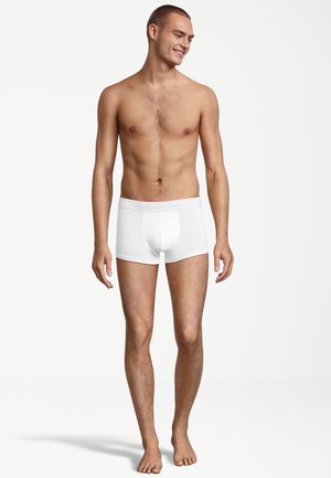 Weiße Baumwoll-Boxershorts mit eng anliegender Passform, versehen mit einem weichen Bund und ohne sichtbare Muster. Das Modell steht barfuß mit einem neutralen Ausdruck.