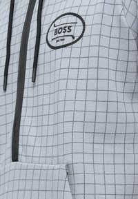 Grå hettejakke med glidelås og rutenettmønster, tekstur av stoff, svarte snorer og en sirkulær "BOSS"-logo på brystet. Frontlomme inkludert.