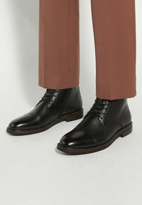 Black leather ankle boots with a rounded toe, featuring lace-up design, subtle texture, and brown sole accents. Styled with tapered brown pants.