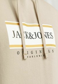 Bež dukserica s vezicom, s velikim prednjim grafičkim prikazom "JACK&JONES" u crnoj i žutoj boji, od teksturiranog pamučnog materijala.