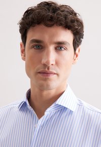 Jeune homme aux cheveux noirs bouclés et yeux bleus portant une chemise habillée rayée bleue et blanche, faisant face à l'avant avec une expression neutre.