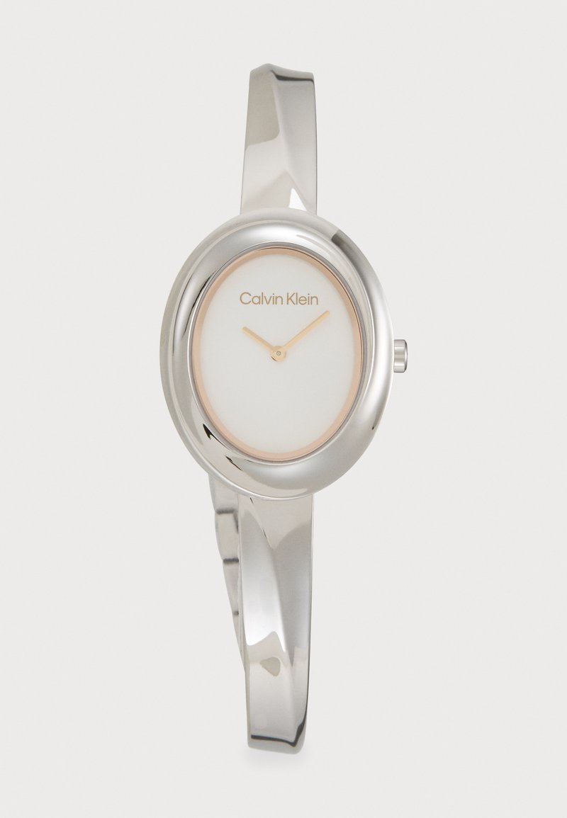 Montre en métal argenté avec un cadran ovale, un fond blanc et des aiguilles minimalistes en ton or. Dotée d'un bracelet de style élégant.