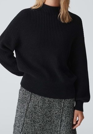 Femme portant un pull en maille côtelée noir et une jupe à motifs noir et blanc sur un fond clair uni.