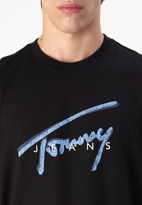 Černé tričko s modro-bílým grafickým designem, na němž je stylizovaný nápis "Tommy" a pod ním ve velkých písmenech "JEANS".