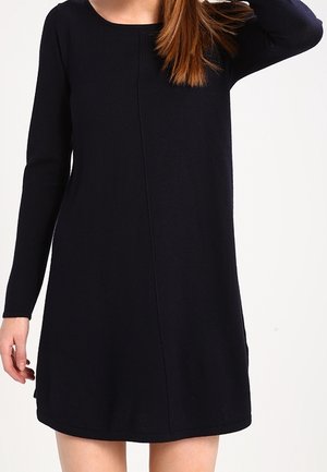 Kvinde iført en langærmet, enkel sort A-formet kjole med rund halsudskæring mod en ensfarvet baggrund.