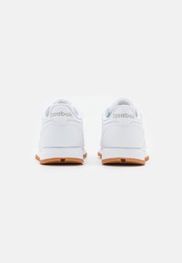 Baskets blanches avec un dessus en synthétique lisse, un col rembourré et une semelle en caoutchouc orange. Présente un logo Reebok sur le talon.