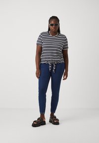 Camisa de manga corta a rayas con nudos en el frente, en azul marino y blanco, combinada con jeans oscuros ajustados y sandalias negras gruesas con tachuelas.