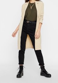 Beige dlouhý blazer přes tmavě olivový žebrovaný top, černé vysoké džíny a robustní černé šněrovací boty. Součástí jsou zlaté vrstvené náhrdelníky.