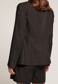 Černý lněný blazer s přizpůsobeným střihem, který má na zadní straně jediný rozventilovaný otvor a čisté linie. Látka má mírnou texturu.