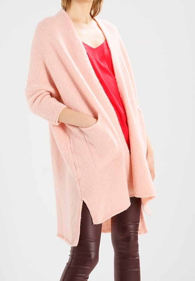 Light pink oversized cardigan with a soft texture, open front, long sleeves, and pockets. Worn over a red top and brown fitted pants.