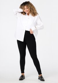 Vrouw met krullend haar in zwarte broek en een losse witte blouse met knopen, glimlachend met één hand achter haar hoofd.