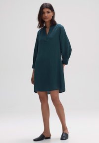 Tealfarbenes, knielanges Kleid mit lockerem Schnitt, V-Ausschnitt und langen, gerafften Ärmeln. Hergestellt aus strukturiertem Stoff und kombiniert mit schwarzen Slippers.