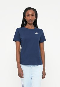 Námornícky modré bavlnené tričko s okrúhlym výstrihom, krátkymi rukávmi a bielym logom Nike na hrudi. Nosené s svetlomodrými nohavicami.