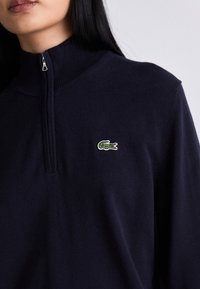 Žena u tamnoplavom puloveru s četvrtastim zipom, s malim zelenim krokodilom na lijevoj strani prsnog dijela.