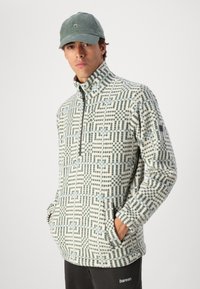 Fleece half-zip pullover in crème met een blauw geometrisch patroon, met een hoge kraag en voorzakken. Draagt een groene pet.
