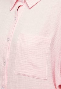 Chemise rose clair texturée à boutons avec une poche poitrine et un col standard, vue en gros plan.