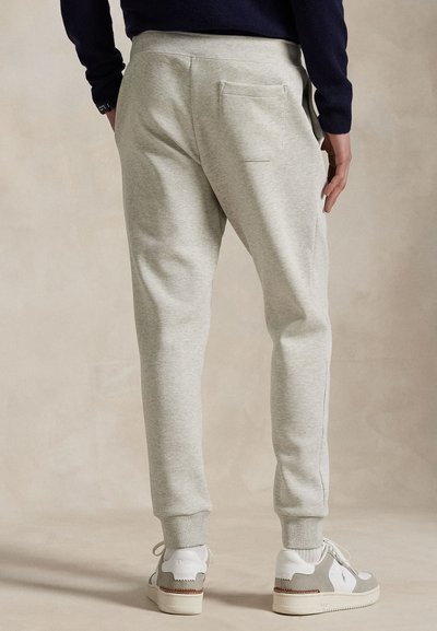 Gaiši pelēkas sporta bikses ar elastīgiem galiem un jostiņu, tām ir aizmugurējā kabata un mīksta tekstūra, valkātas kopā ar baltām un pelēkām sporta kurpēm.