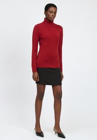 Maglione a collo alto rosso in morbido tessuto lavorato a maglia, design aderente, abbinato a una mini gonna nera e mule con tacco neri.