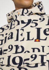 Fleece-Pullover in Creme mit auffälligem marineblauem Text und Zahlen, Halb-Reißverschluss, Vordertasche und kleinem, farbigem Logo auf der Brust.