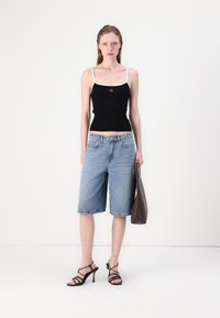 Svart ribbenet tanktop med hvite stropper, blå denim bermudashorts, og svarte stroppeesko. Grå håndveske holdes i høyre hånd.