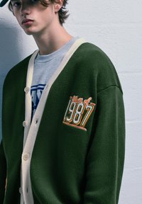 Giovane che indossa un cardigan verde con un patch "Classe del 1987", una maglietta grigia chiara e un cappellino scuro, in piedi contro un muro chiaro e texturizzato.