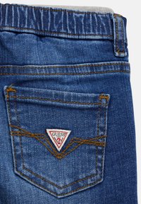 Denim auduma džinsi zilā krāsā, ar aizmugurējo kabatu, kurai ir sarkans un balts trīsstūra logo un brūna šuvju akcenti.