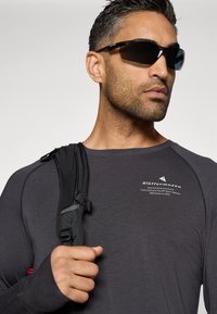 Γκρι μακρυμάνικο πουκάμισο με λογότυπο στο στήθος, συνοδευόμενο από μαύρα γυαλιά ηλίου και ένα μαύρο λουρί σακιδίου στον ώμο.