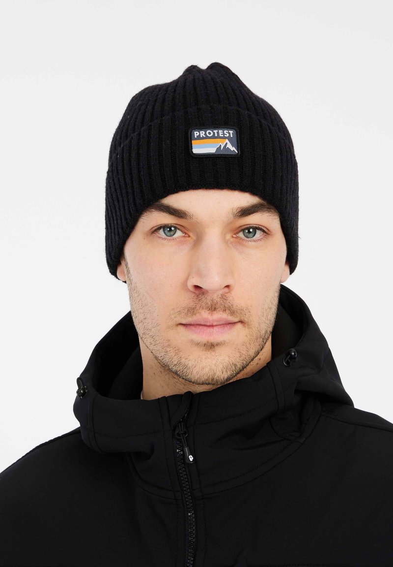 Berretto a coste nero con una piccola patch rettangolare che mostra montagne e il testo "PROTEST." Indossato con una giacca nera con zip.