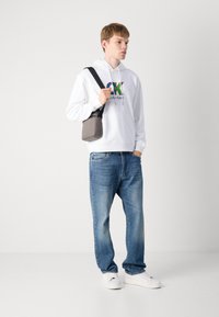 Sweatshirt à capuche blanche avec logo coloré, jean bleu en denim et baskets blanches. Sac bandoulière gris avec sangle réglable.