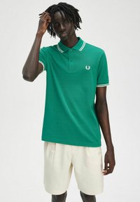 Chemise polo verte avec des rayures blanches sur le col et les manches, arborant un petit logo brodé sur la poitrine, associée à un short crème.
