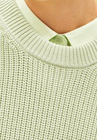 Lähellä otettu kuva vaaleanvihreästä neulotyöstä, joka on kerroksittain vaaleanvihreän kauluspaitapuseron päällä henkilön kaulalla ja harteilla.